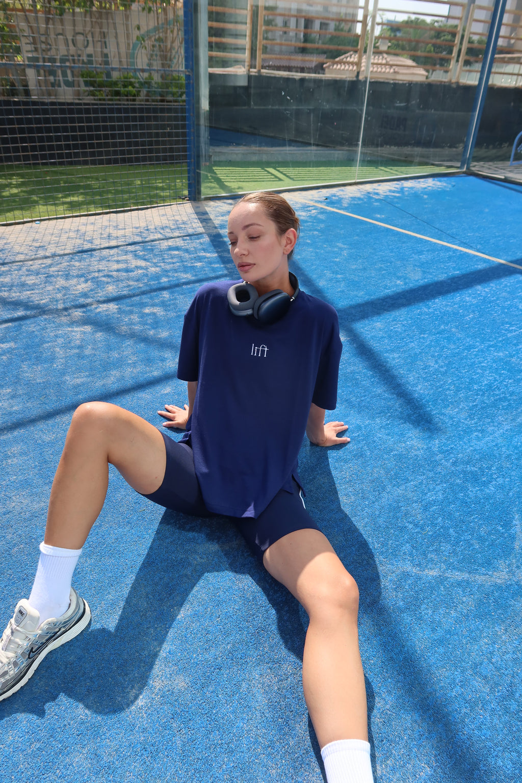 Model sits on a blue tennis court wearing a navy blue t-shirt with white embroidered 'lift' logo on the chest, matching navy shorts, white socks, and athletic shoes, with black headphones around her neck.