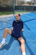Model sits on a blue tennis court wearing a navy blue t-shirt with white embroidered 'lift' logo on the chest, matching navy shorts, white socks, and athletic shoes, with black headphones around her neck.