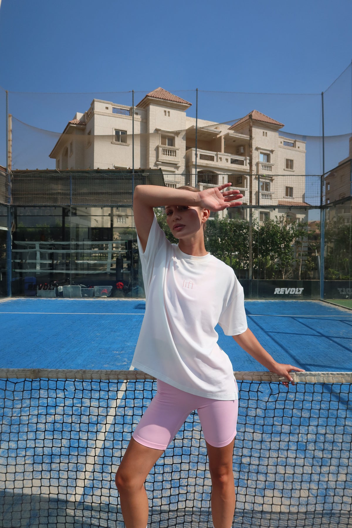 A woman posing on a blue tennis court wearing a white oversized t-shirt with a small chest logo, light pink fitted shorts, and sunglasses. The background shows a tennis net, court, and a multi-story building under a clear blue sky in Egyptian local activewear brand.
